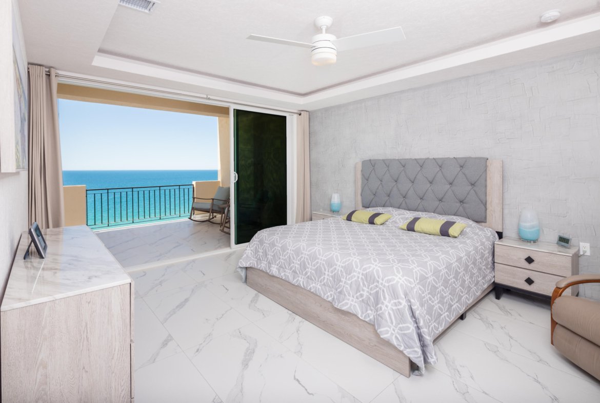 Bright ocean-view bedroom with a sliding door to a balcony, a queen bed with a tufted gray headboard, and marble floors.