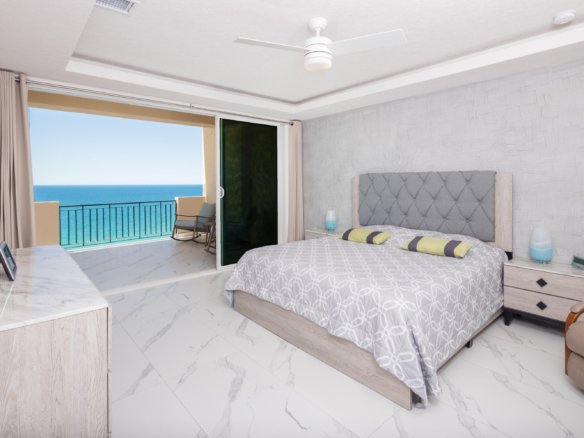 Bright ocean-view bedroom with a sliding door to a balcony, a queen bed with a tufted gray headboard, and marble floors.