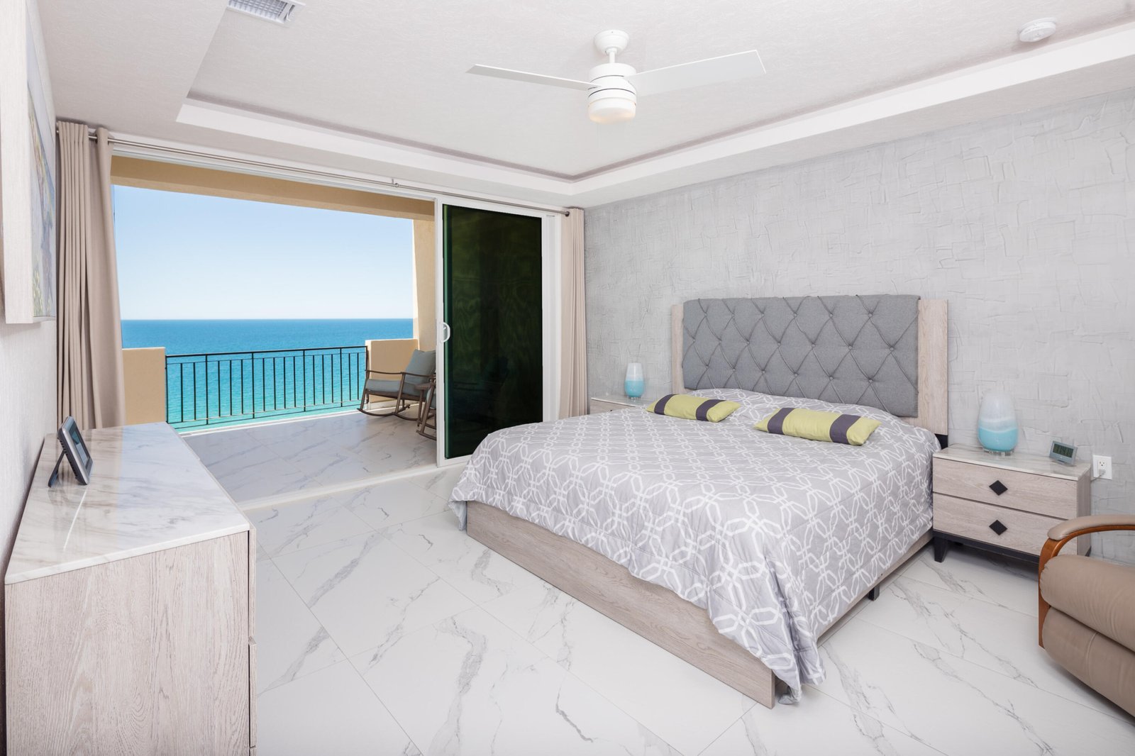 Bright ocean-view bedroom with a sliding door to a balcony, a queen bed with a tufted gray headboard, and marble floors.