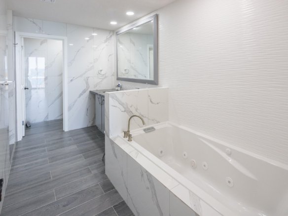 Contemporary bathroom with a built-in marble tub, grey wood-look floor, marble walls, and a large framed mirror above the vanity.