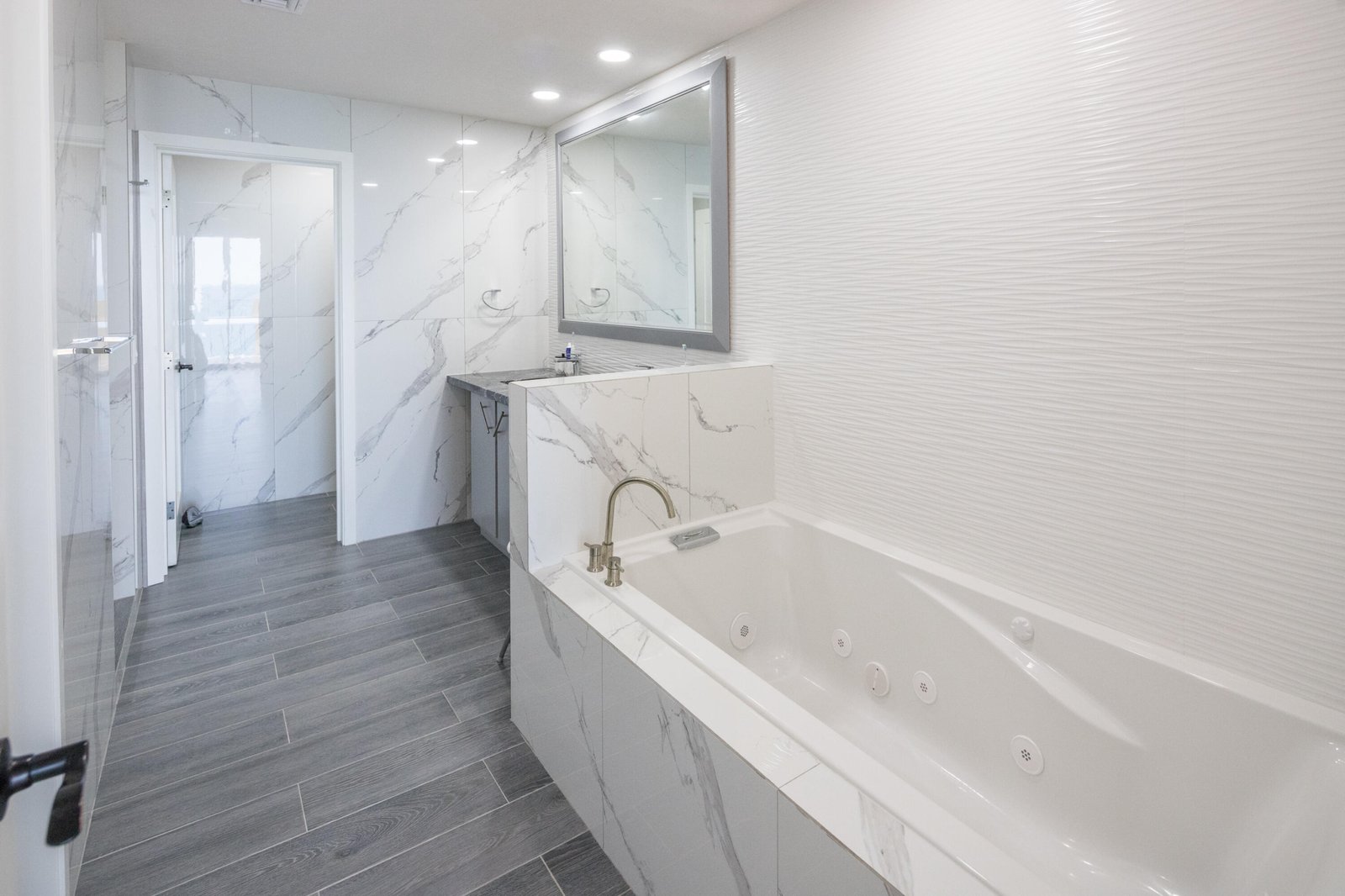 Contemporary bathroom with a built-in marble tub, grey wood-look floor, marble walls, and a large framed mirror above the vanity.