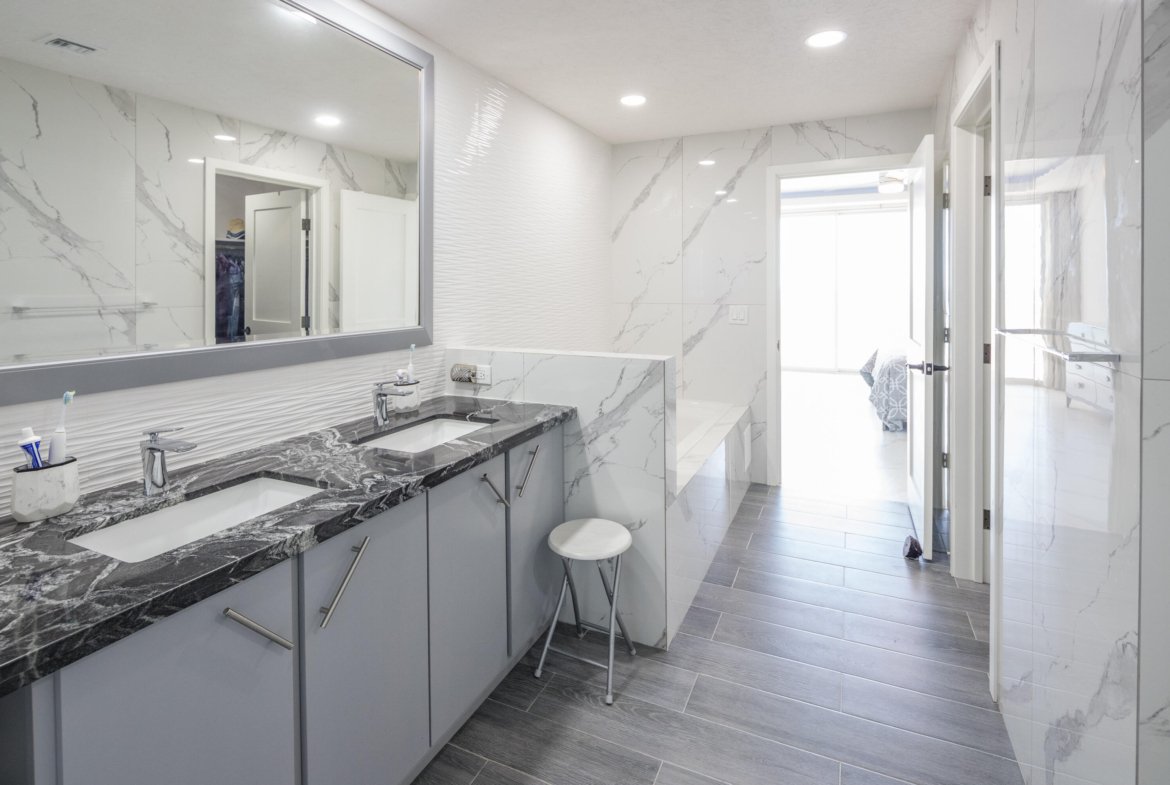 Modern bathroom vanity with gray cabinets, dark marble countertop, dual sinks, and a large framed mirror reflecting a bright, marble-tiled wall.”] ,