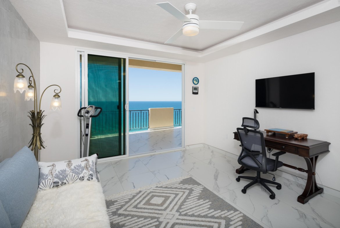 Bright living/office area with a wooden desk and rolling chair facing a wall-mounted TV, and a sliding glass door to a balcony with an ocean view.