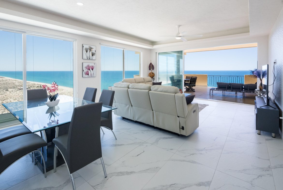 Bright coastal living room with a glass dining table, gray chairs, and a white sofa, overlooking the ocean through expansive windows.