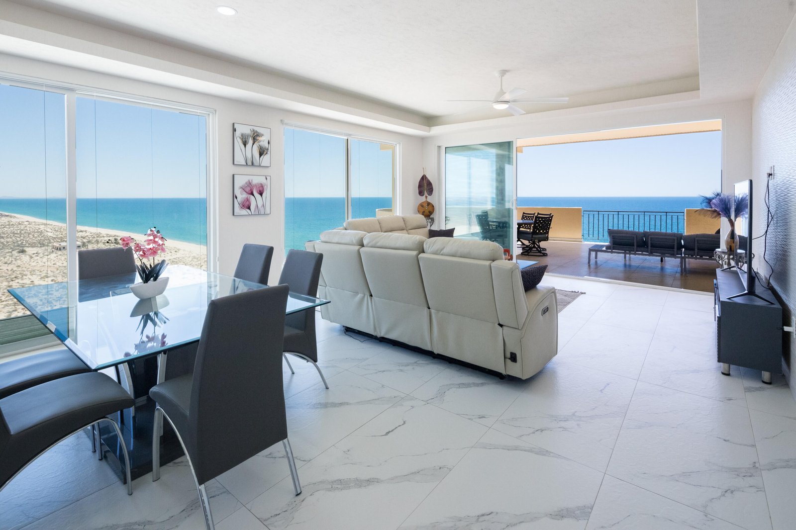 Bright coastal living room with a glass dining table, gray chairs, and a white sofa, overlooking the ocean through expansive windows.