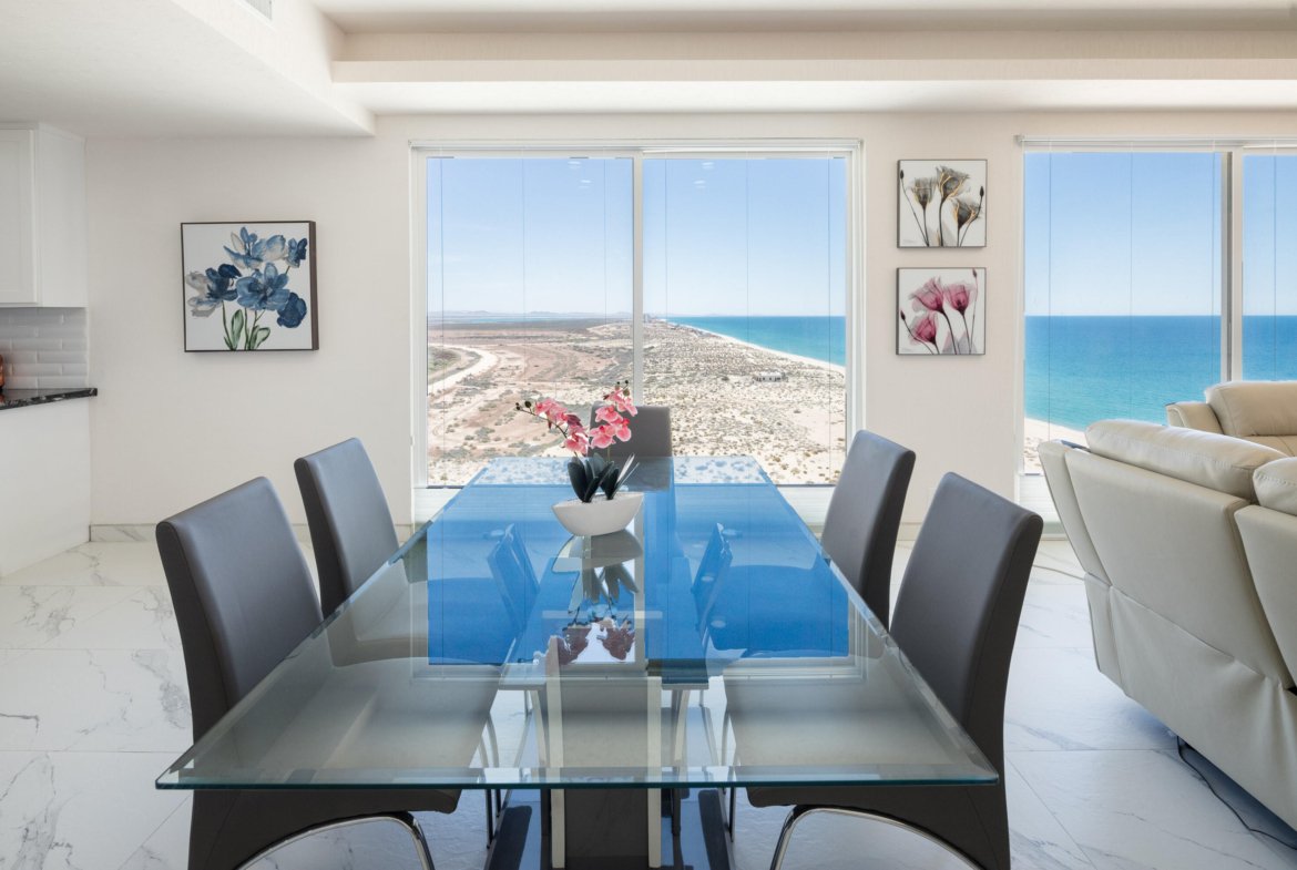 Dining room with a glass-top table and gray chairs, large windows reveal a desert landscape and blue ocean beyond.