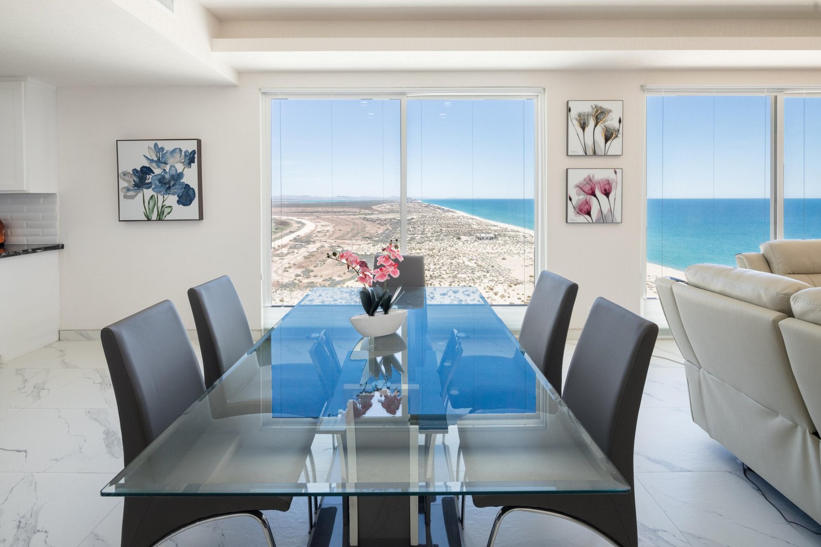 Dining room with a glass-top table and gray chairs, large windows reveal a desert landscape and blue ocean beyond.