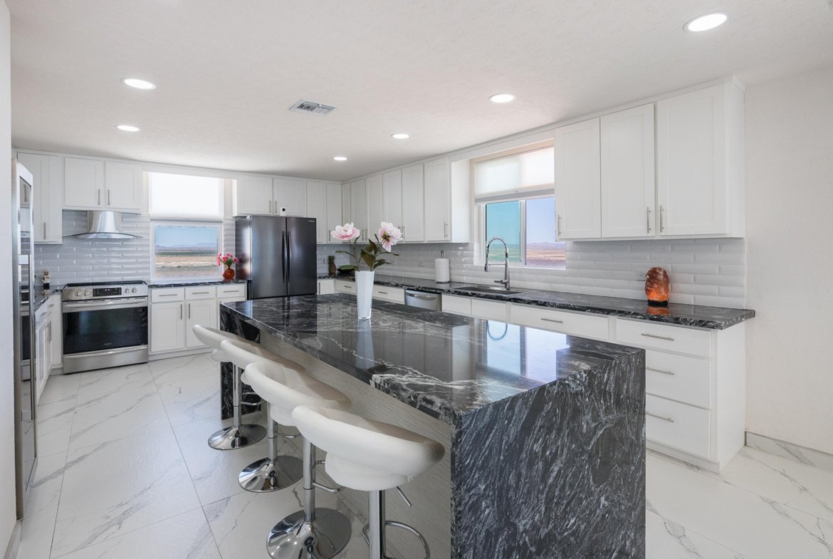 Bright white kitchen with a dark marble island, marble floors, and stainless steel appliances including a large refrigerator and stove. Smooth white cabinets line the walls, with a window over the sink.