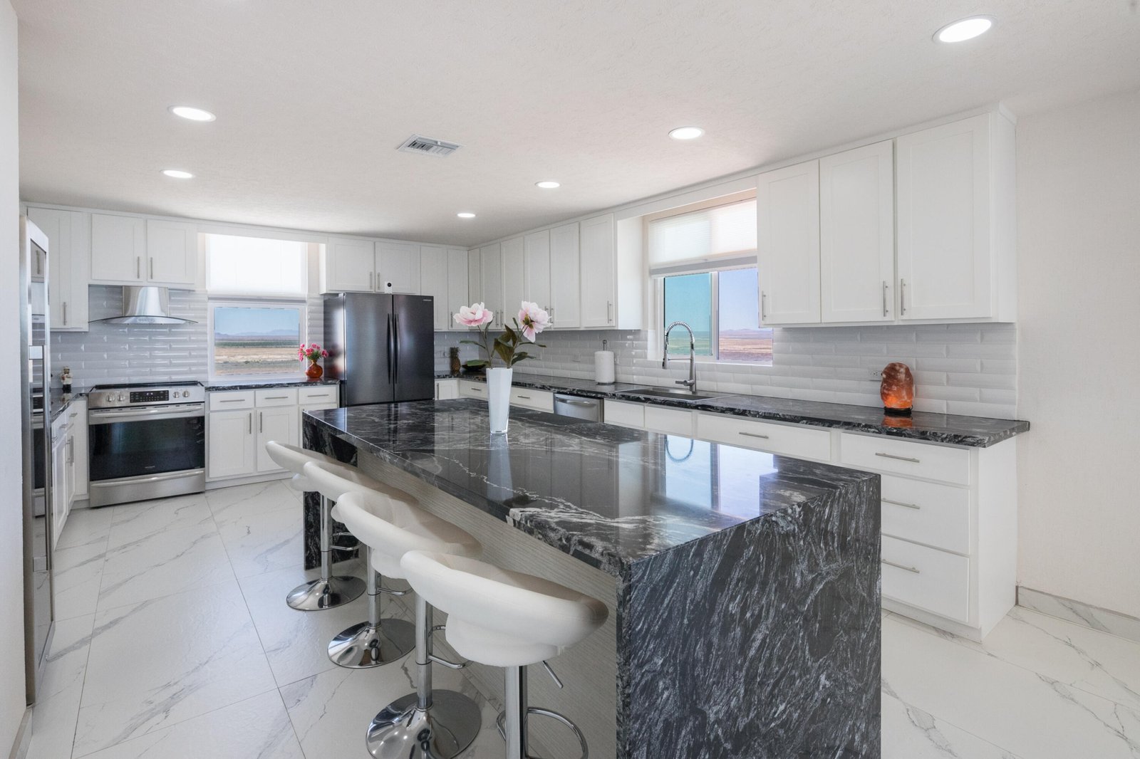 Bright white kitchen with a dark marble island, marble floors, and stainless steel appliances including a large refrigerator and stove. Smooth white cabinets line the walls, with a window over the sink.